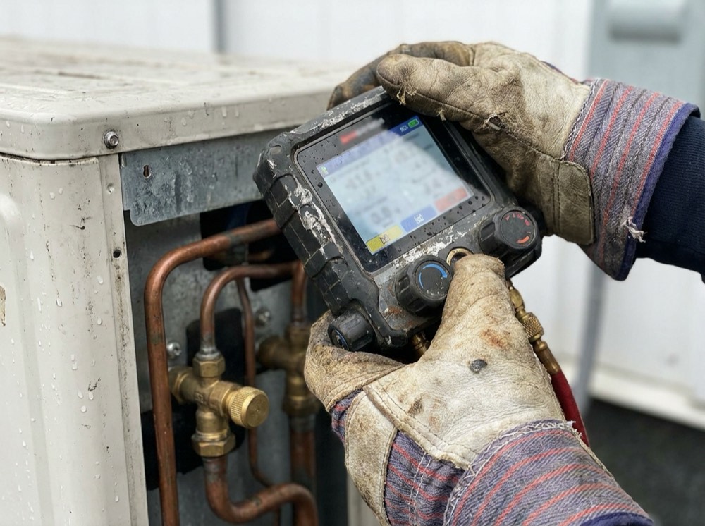 Gloved hands holding a rugged handheld digital manifold gauge over a mini-split outdoor unit